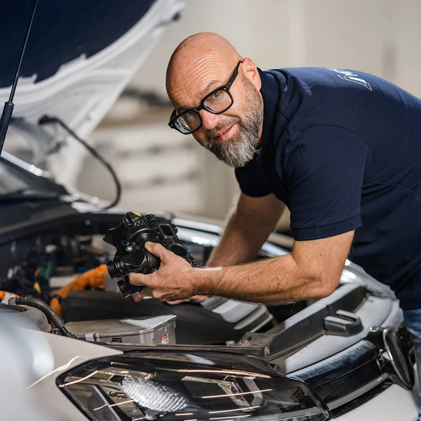 The well-known German mechanic Andreas Raulfs leans into the engine compartment of a car and holds a MEYLE ORIGINAl part.