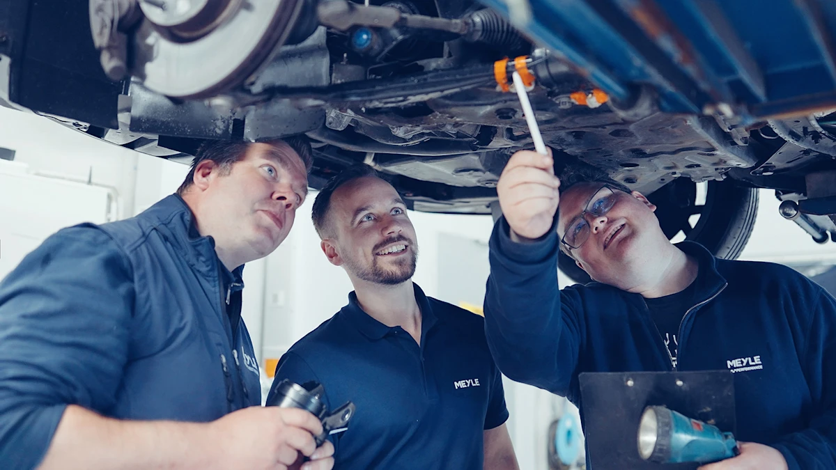 Three people stand on a lifting platform under a car and analyze a car spare part.