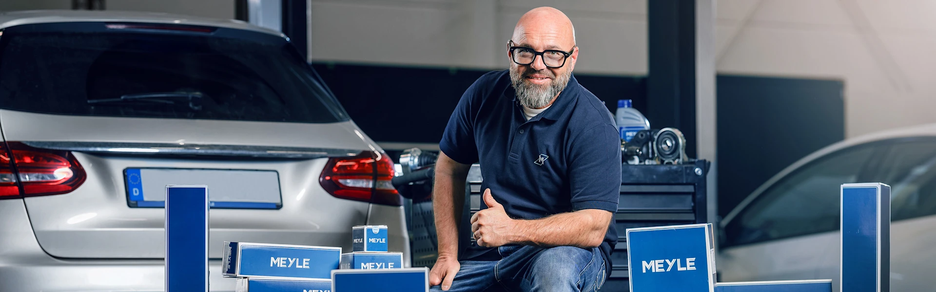 Person sits on a tire in the workshop, gives a thumbs-up to the camera and smiles. Many MEYLE boxes are stacked in front of him. There is a car in the background.