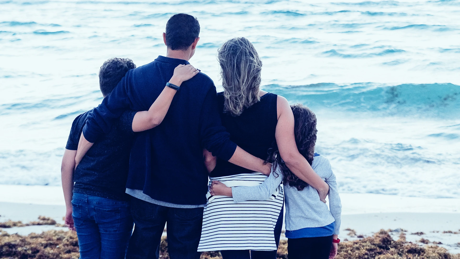 A family with 2 children stand with their backs to the camera and the sea in front of them.