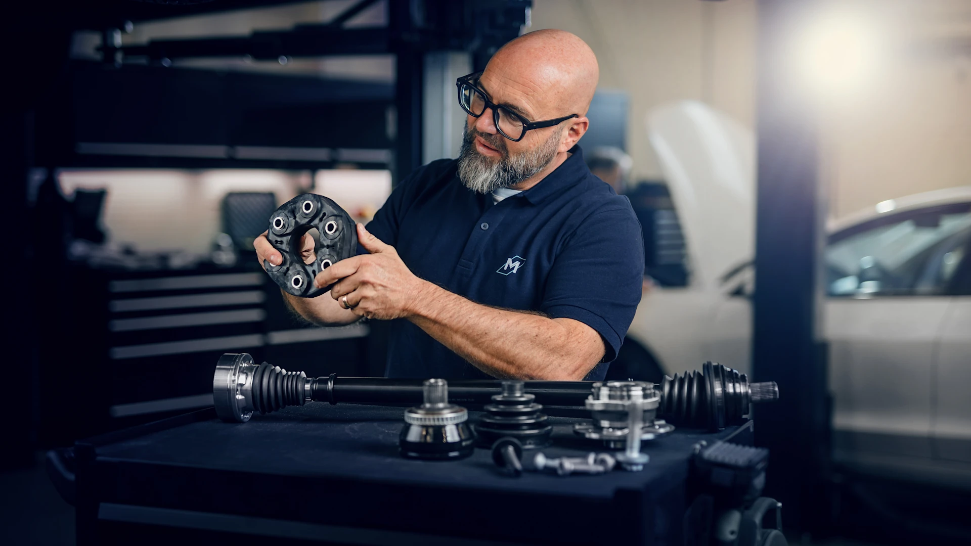 Photo of Andreas Raulfs standing in a workshop. In front of him is a tool trolley on which many MEYLE drive parts are lying. He is holding a MEYLE flex disc in his hand.