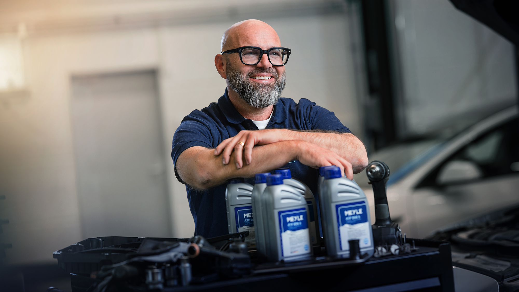The well-known German mechanic Andreas Raulfs is in a workshop and leans on MEYLE transmission oil bottles with a smile.