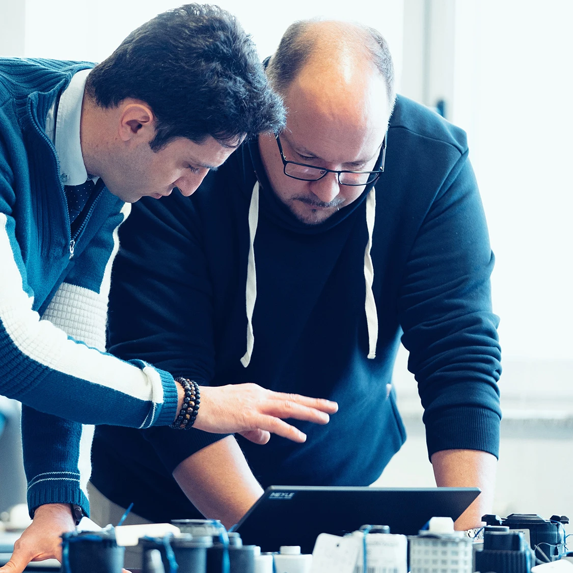 Two people bend over a laptop and show each other something. There are several items on the table in front of them.