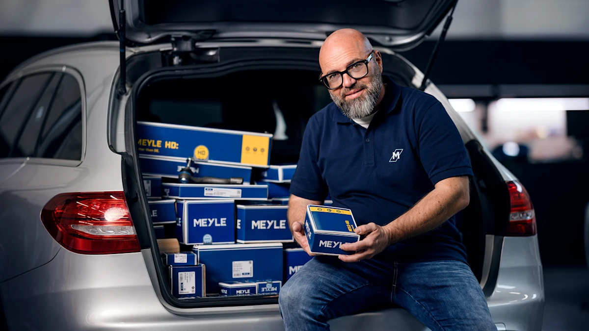 Person sitting on the edge of an open trunk in a workshop. The trunk is filled with MEYLE boxes. The person is holding a MEYLE HD box in their hands.