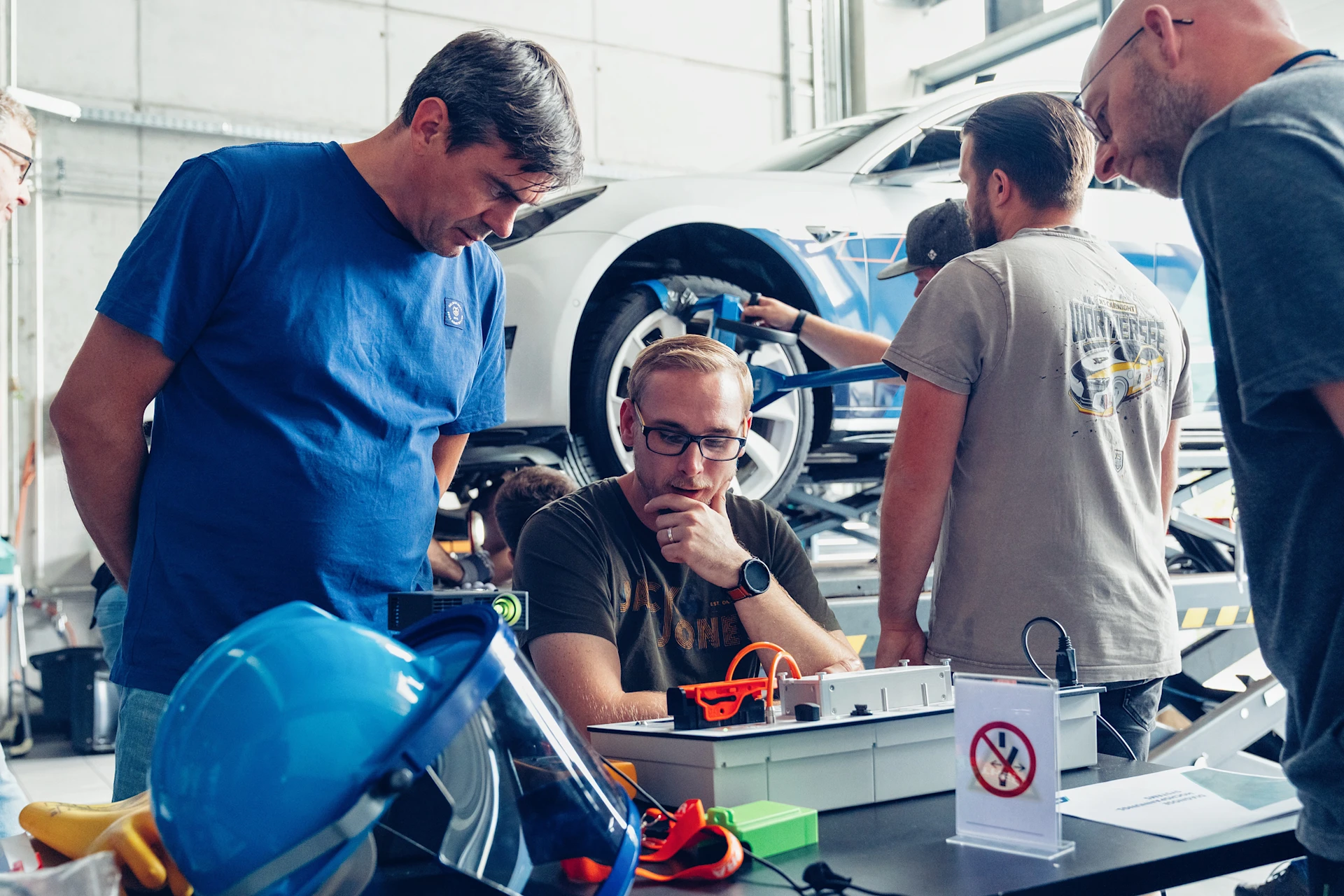 EV Training in MEYLE Werkstatt. Teilnehmer sitzt am Tisch, andere Teilnehmer stehen um ihn herum und sie schauen sich eine Elektroauto Batterie an.