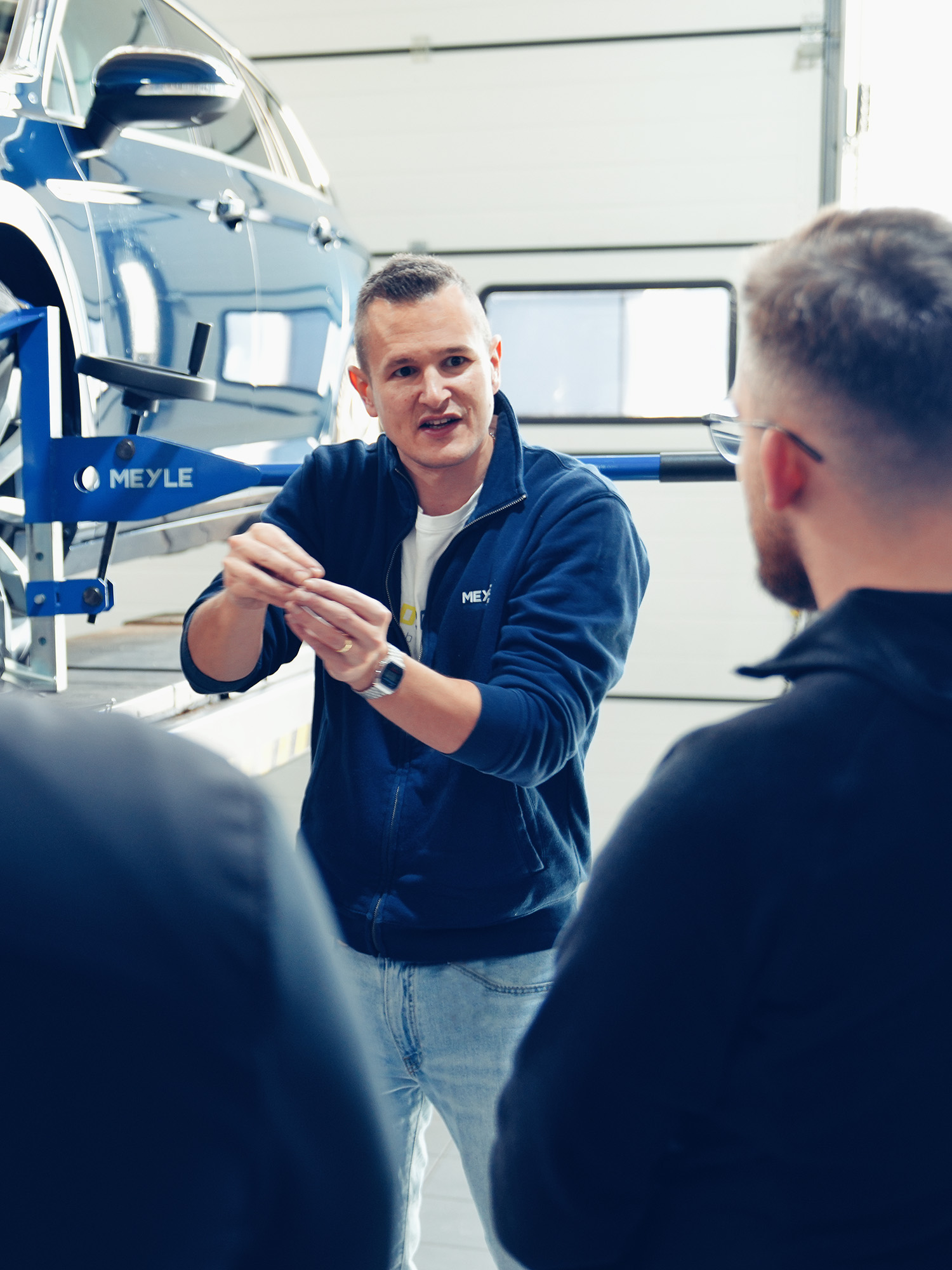 Person explains something to other people on the lifting platform, in the background you can see the MEYLE articulated play tester on the car.