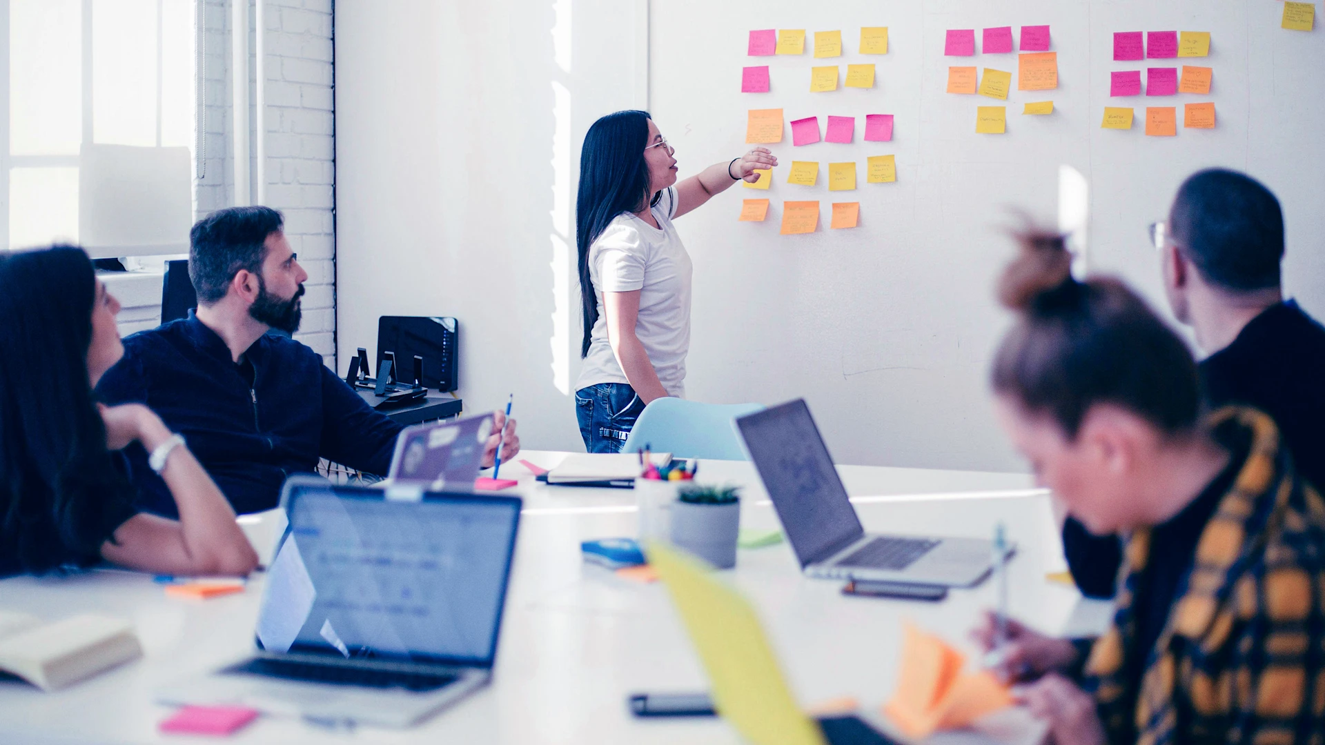 Four people sit at a table and watch a person standing at a whiteboard and sticking sticky notes on it.