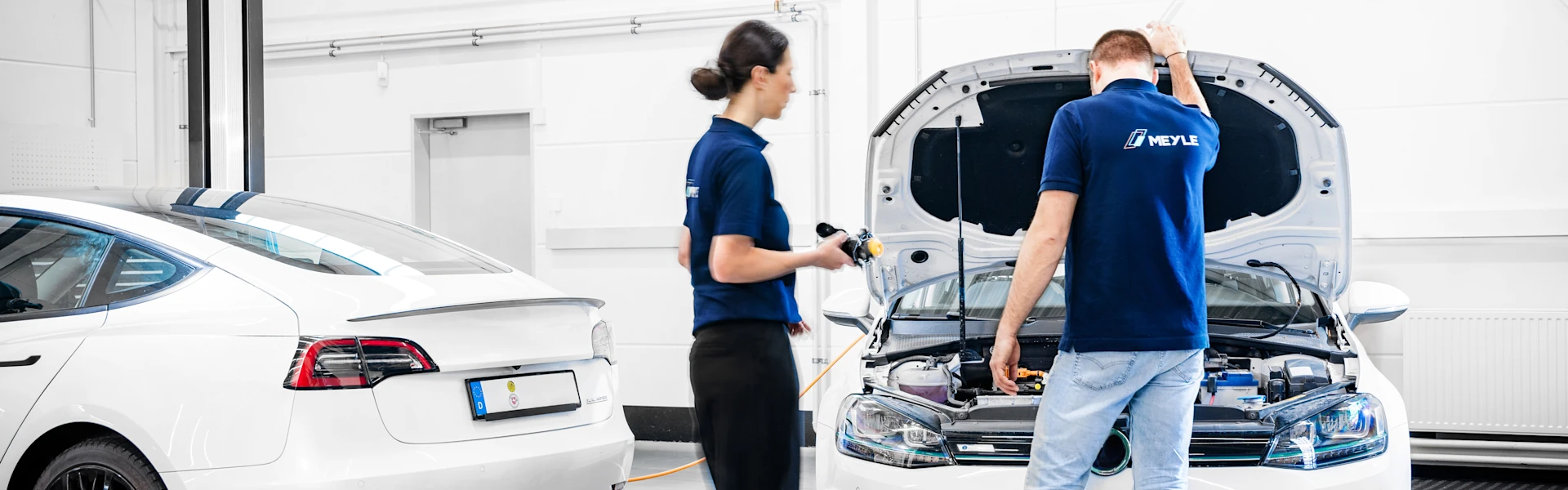 Two people are in a workshop where there are two white electric cars, one of which has the hood open and one of the people is standing in front of the open hood with his back to us.