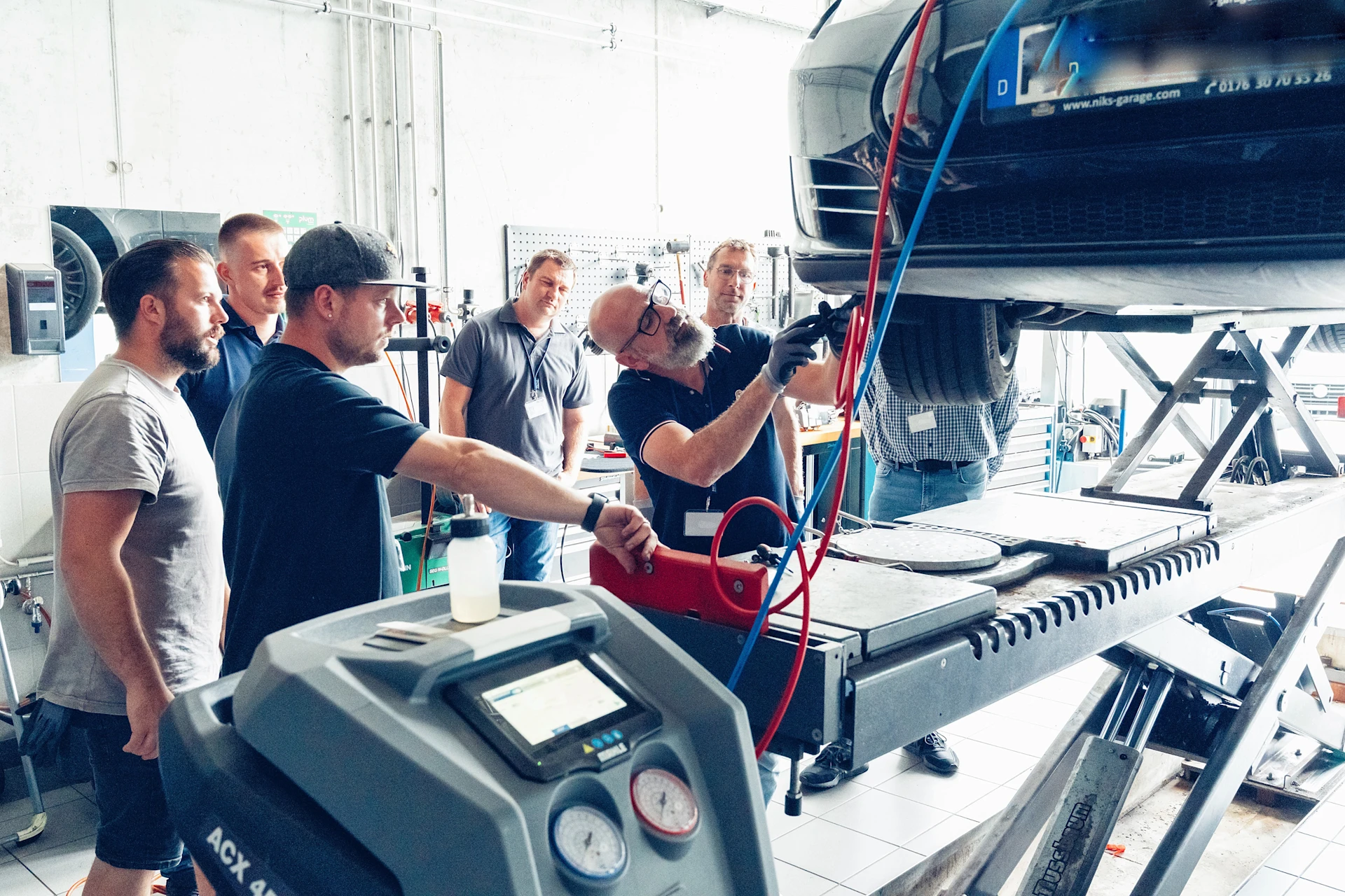 EV training in the MEYLE workshop. Participants stand next to the car, which is on a lifting platform, and work on it.