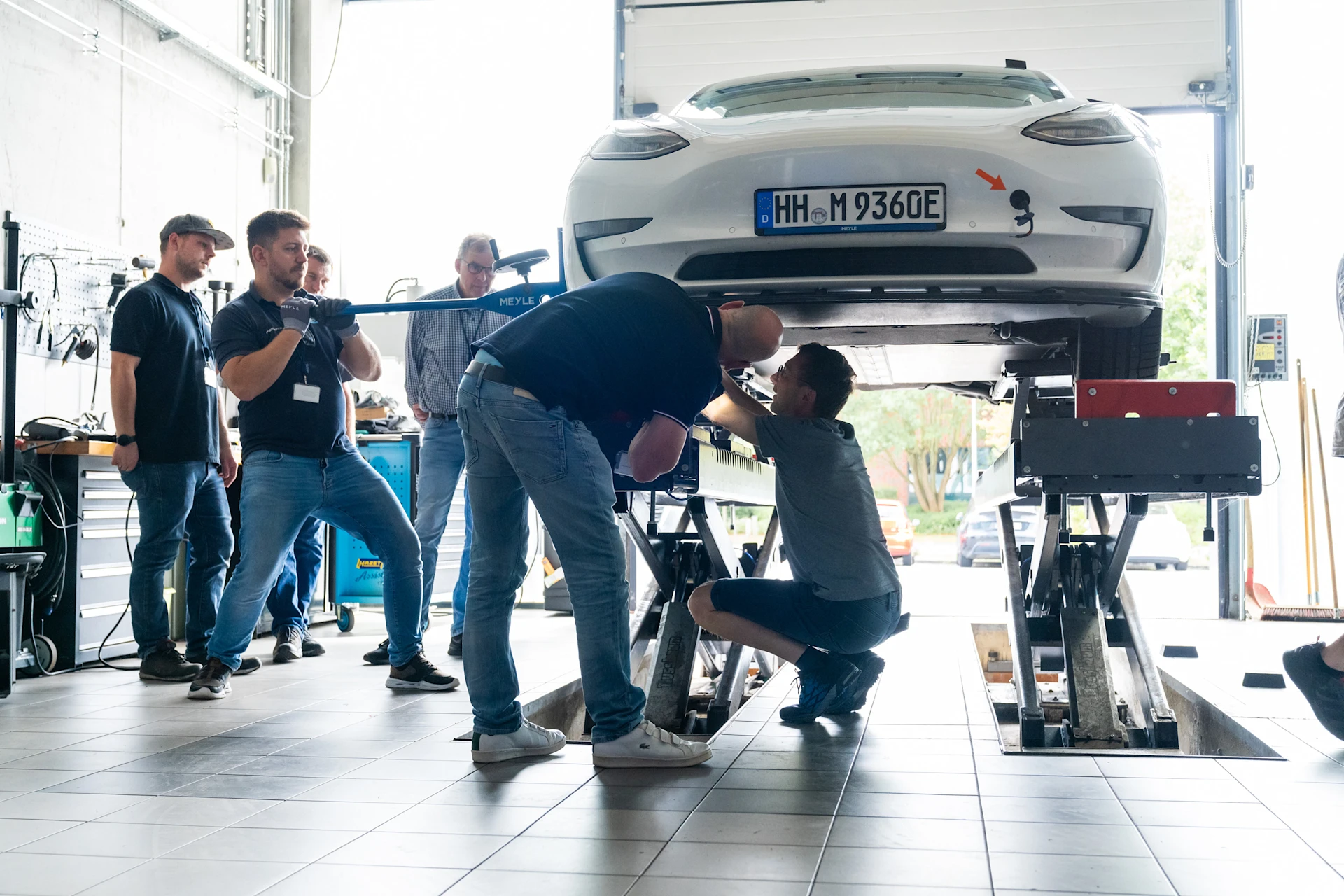 EV Training in der MEYLE Werkstatt. Trainingsteilnehmer stehen an der Hebebühne und arbeiten an einem Elektroauto.