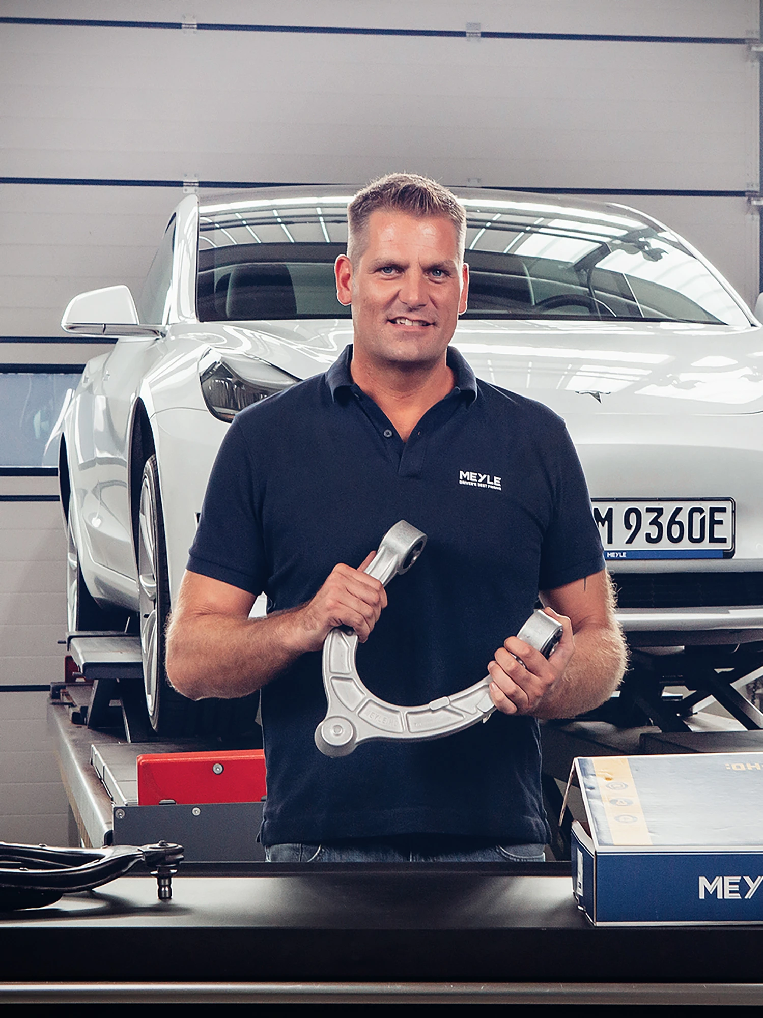 Trainer zeigt MEYLE Querlenker, im Hintergrund ist ein Auto auf einer Hebebühne.