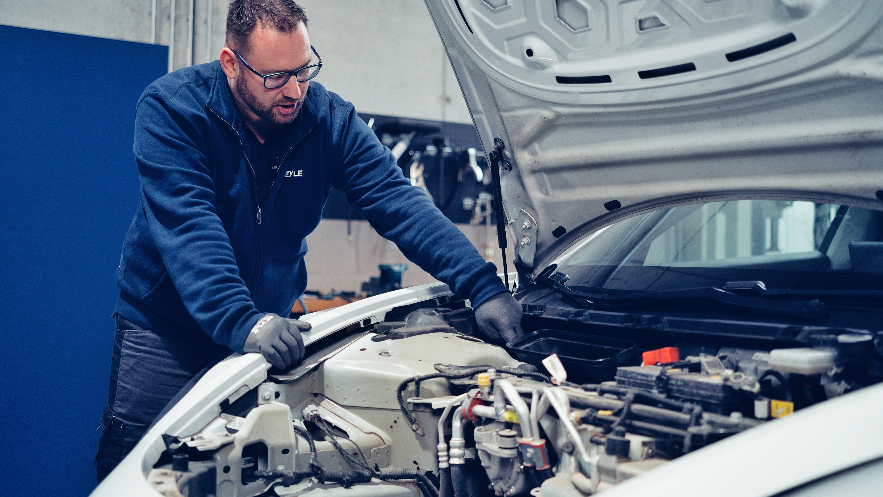Mechanic in high-voltage protective clothing stands at the open engine compartment of an electric vehicle.