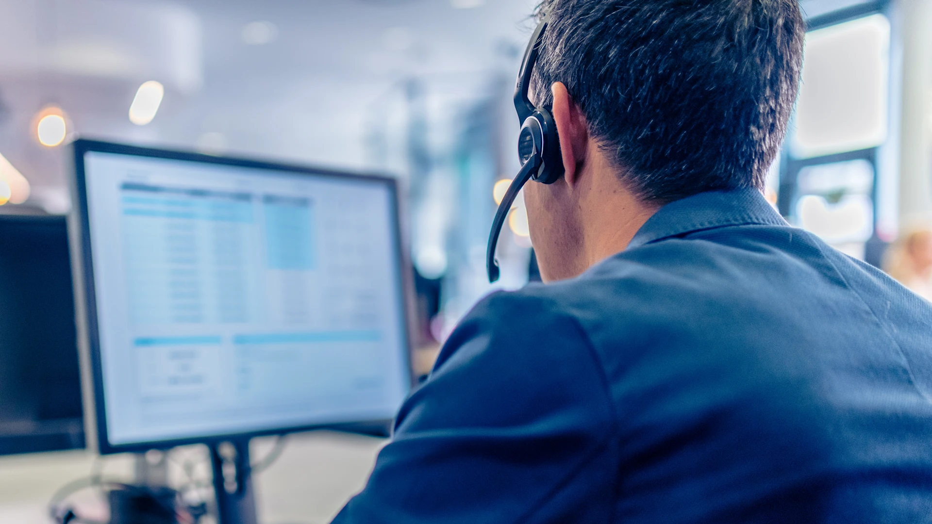 Person von hinten mit Headset, Kopf im Fokus, im Hintergrund ist ein Monitor in der Unschärfe.