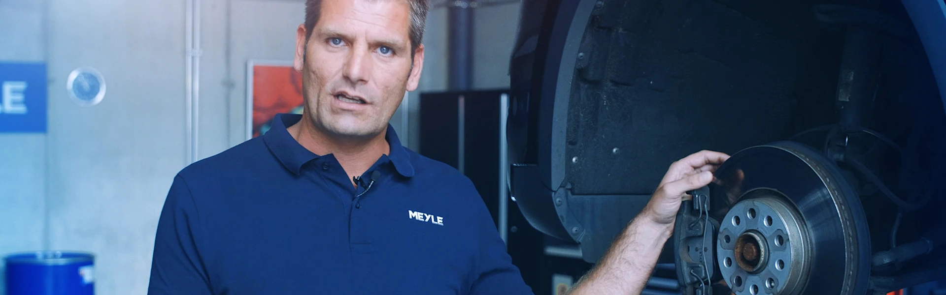 Person steht an einem Auto das auf einer Hebebühne steht und zeigt auf eine verbaute Bremsscheibe mit Bremsbelägen.