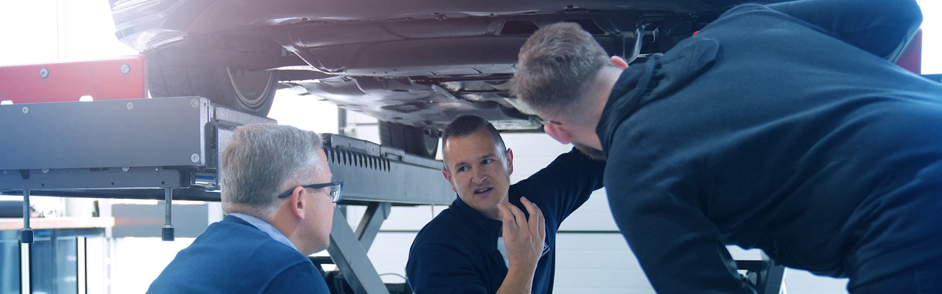 One person explains something to two other people under the car on a lifting platform.