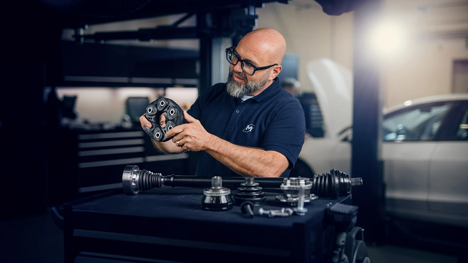 Person standing in front of a tool trolley in a workshop. Several MEYLE drive parts are lying on it. The person is holding a flex disc.