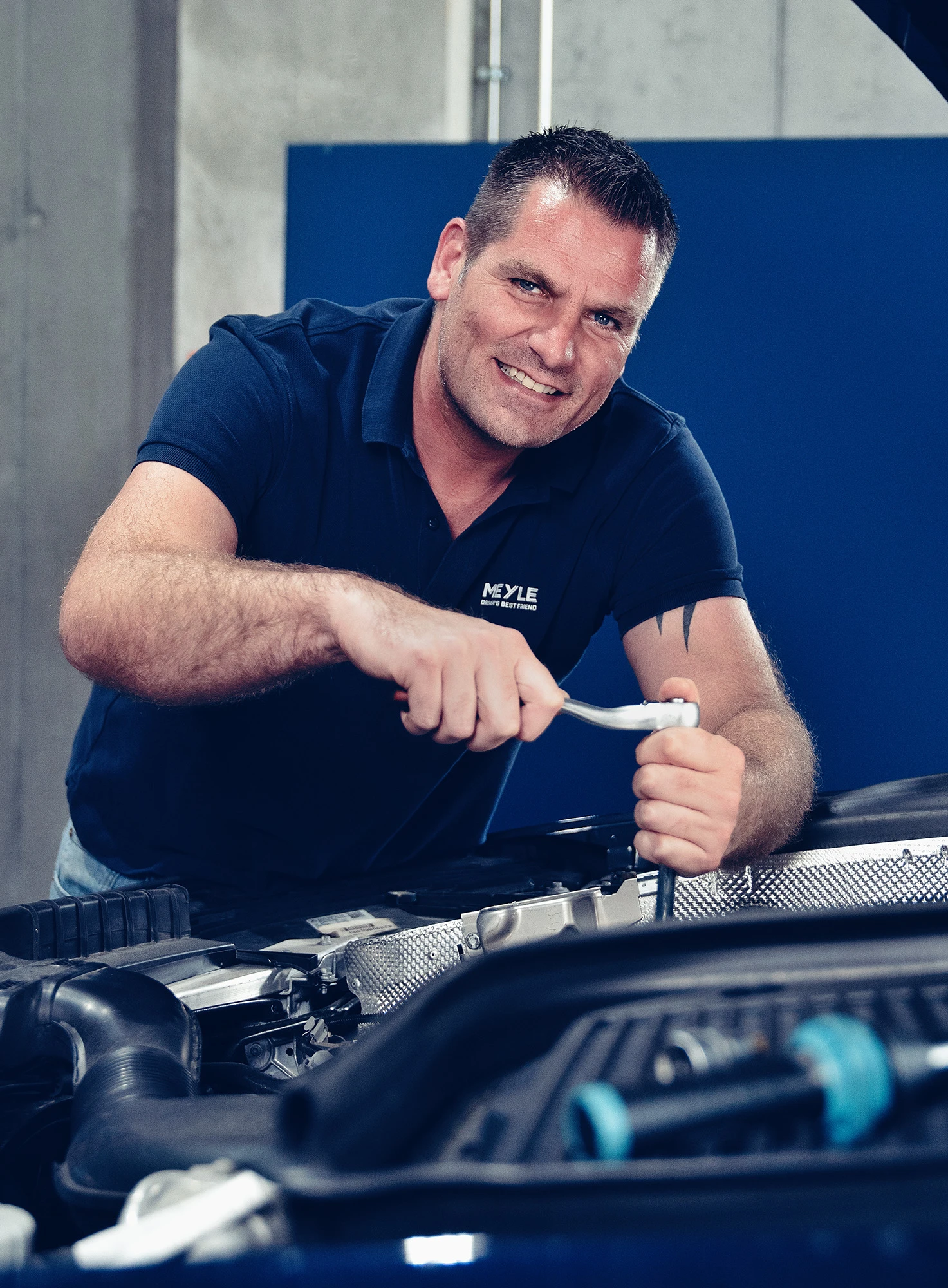 Mechaniker schraubt im Motorraum und lächelt in die Kamera.
