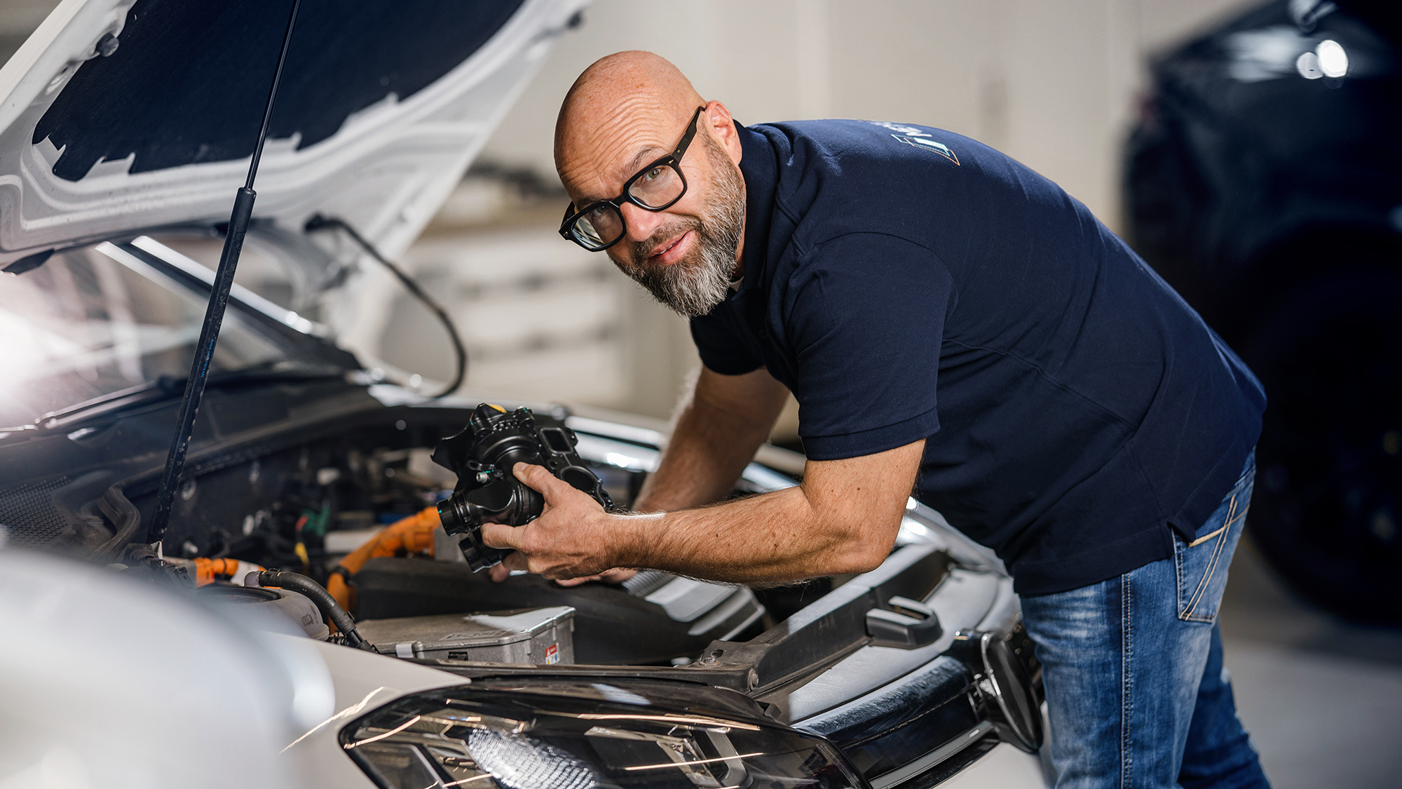 The well-known German mechanic Andreas Raulfs leans into the engine compartment of a car and holds a MEYLE ORIGINAl part.