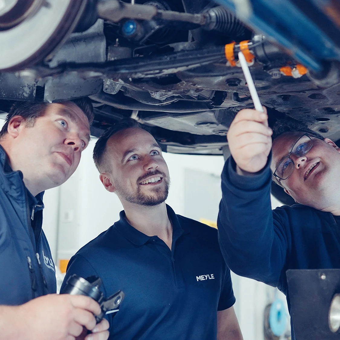 Three people stand under a car on a lifting platform and identify problems.