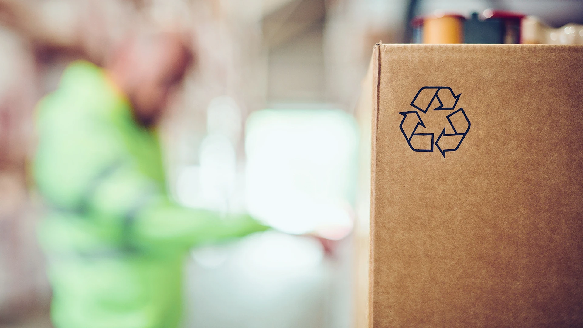 Cardboard with a recycling icon in the background, a person can be seen out of focus.