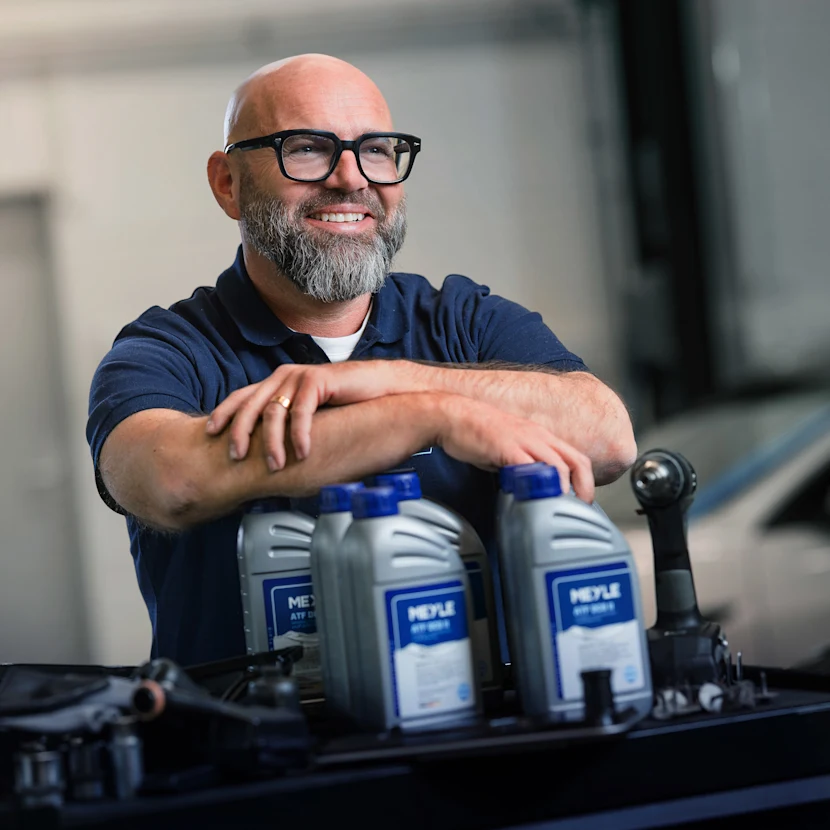 The well-known German mechanic Andreas Raulfs is in a workshop and leans on MEYLE transmission oil bottles with a smile.